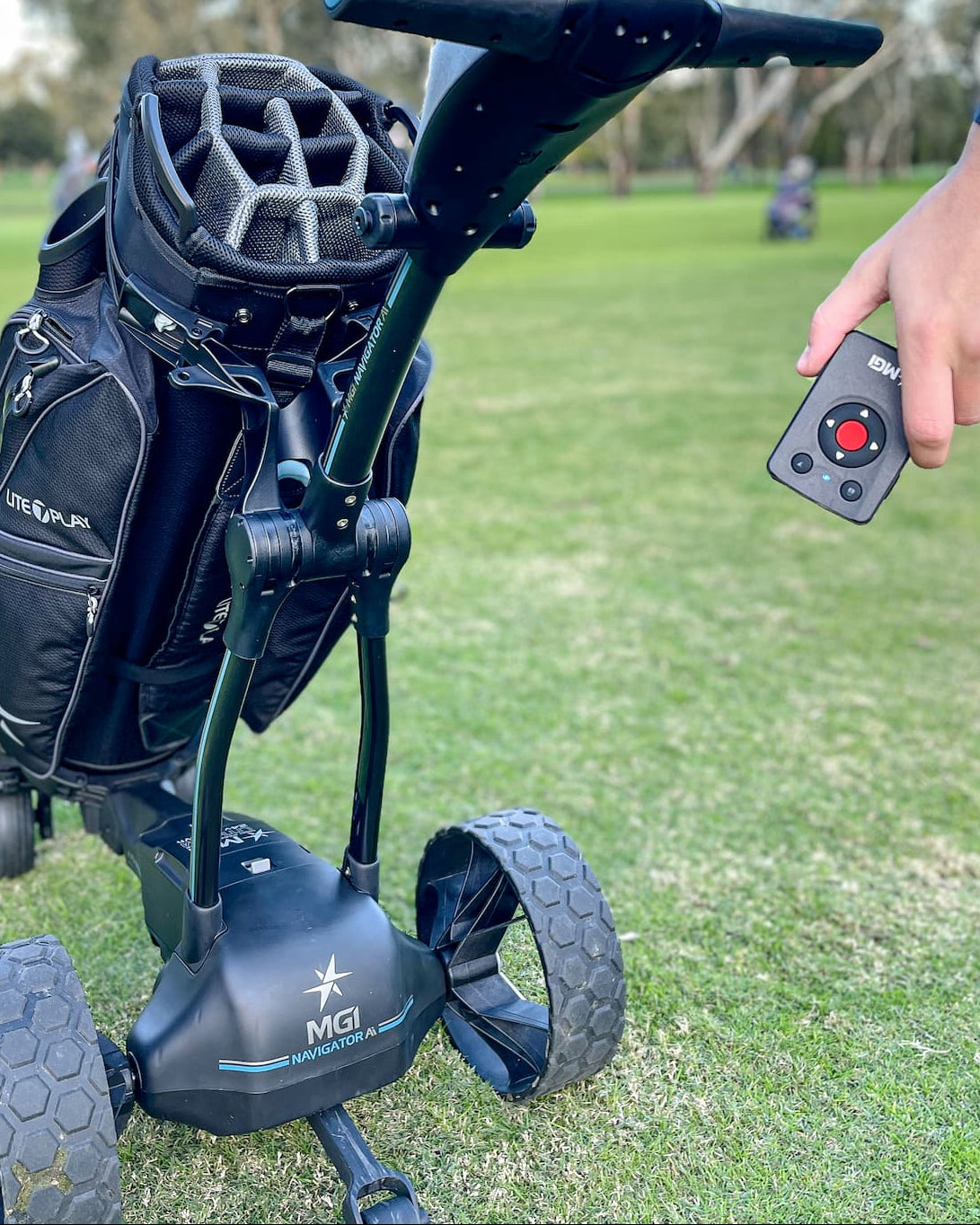 A black golf trolley with a golf bag on a grassy golf course, with a person's hand holding a small remote control.