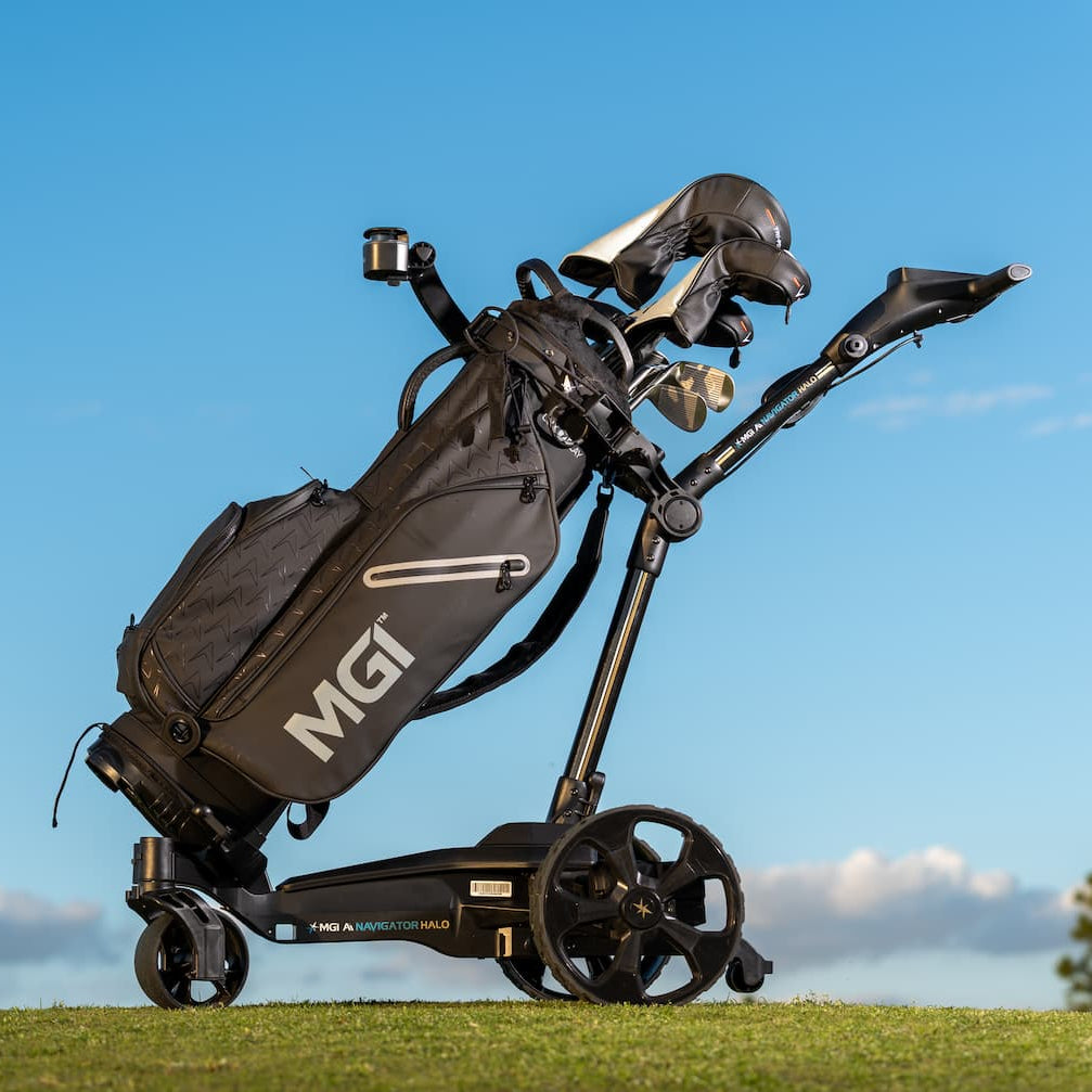 A black MGI golf trolley with a golf bag loaded with clubs, positioned on a grassy golf course under a bright blue sky.