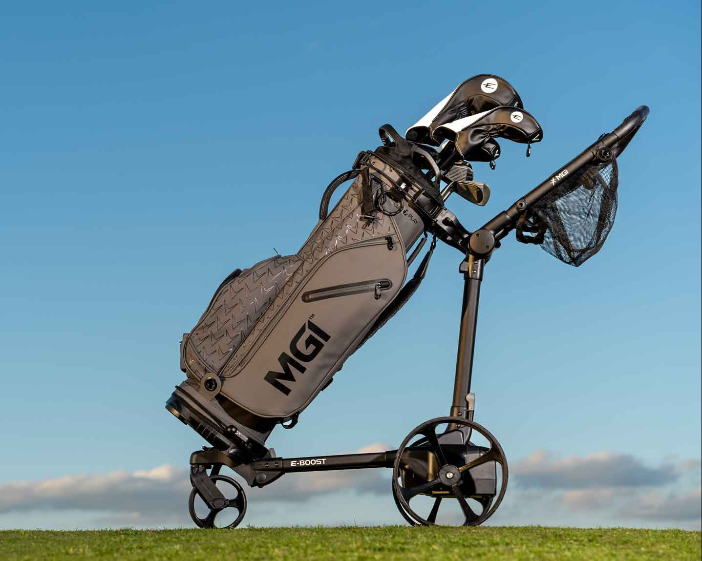 Gray MGI golf bag mounted on a three-wheeled electric golf push cart, shown in side view on grass against a clear blue sky.