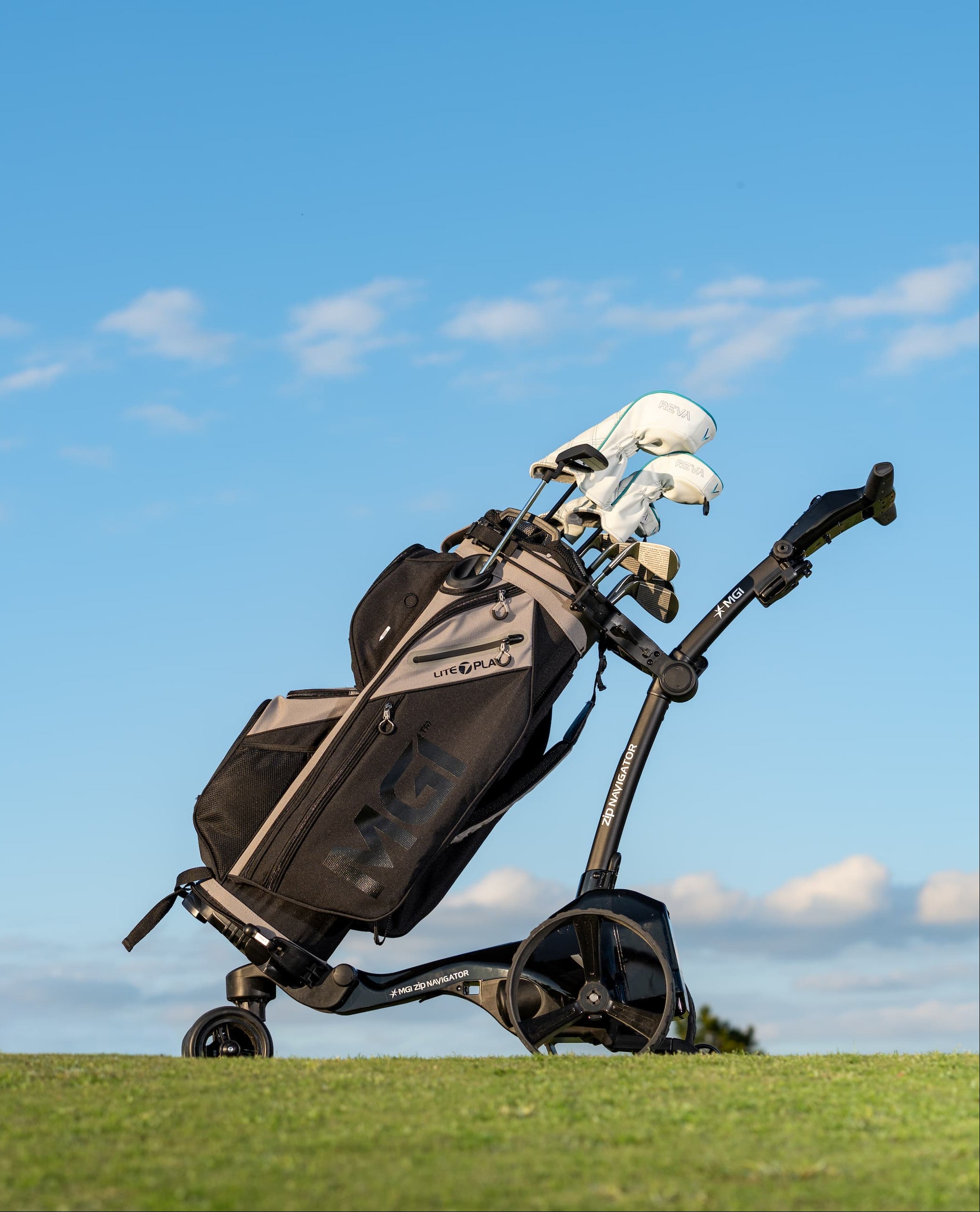 A black and gray golf bag with golf clubs on a golf trolley, set on a grassy field under a bright blue sky with light clouds.