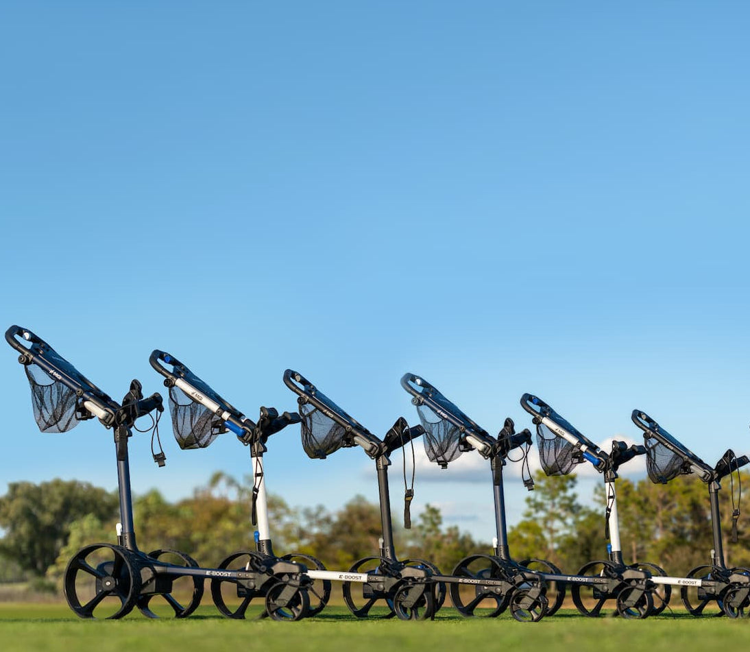 A row of golf trolley carts with net bags lined up on a grassy field under a clear blue sky.