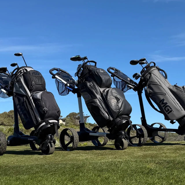 Three golf bags mounted on black three-wheeled golf carts parked on a grassy golf course under a clear blue sky. The bags are dark gray and black, with one bag showing the MGI brand name.