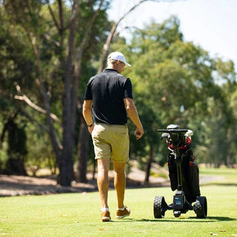 A man on a golf course seen from behind, wearing a black polo shirt, khaki shorts, and a white cap, walking beside a black electric golf trolley with golf clubs.